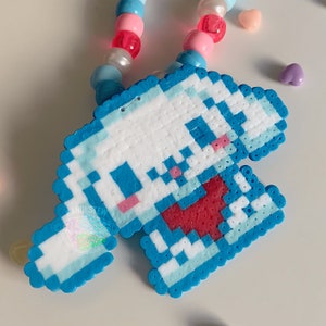 May include: A blue and white pixelated character with a red mouth, made from small plastic beads. The character is attached to a colorful beaded necklace.
