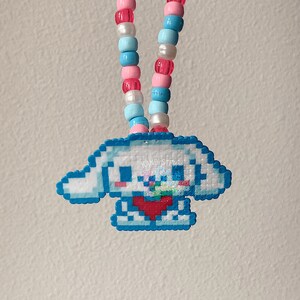 May include: A blue and white pixelated charm of a cartoon dog with a red heart, hanging from a pink, blue, and white beaded necklace.