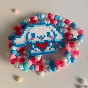 May include: A white and blue kandi bracelet with a pixelated white puppy charm with a red heart. The bracelet is made of colorful beads in pink, blue, and white.