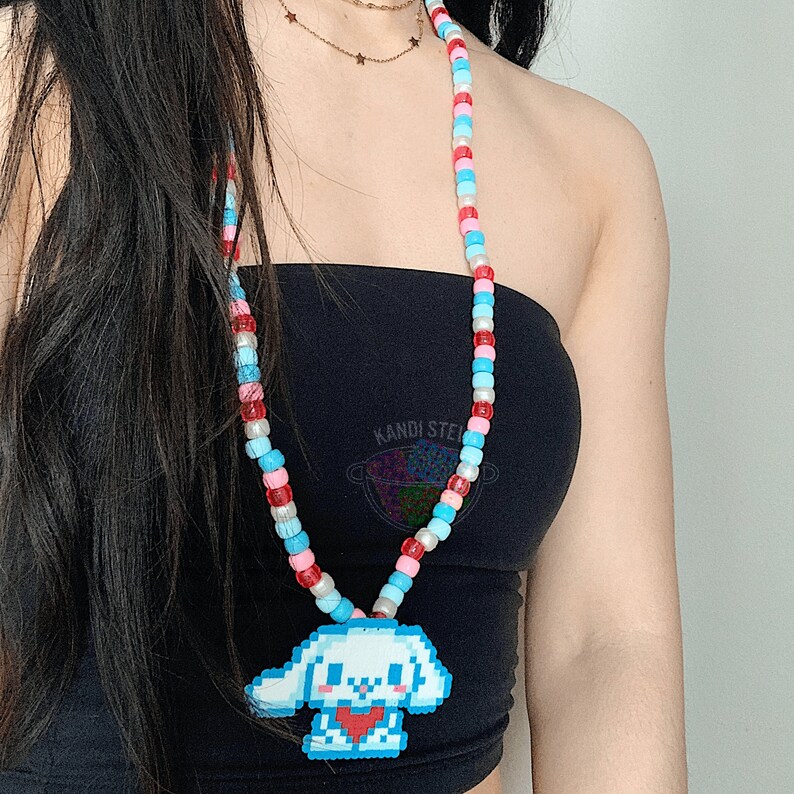 May include: A colorful beaded necklace with a pixelated dog charm. The necklace is made of red, blue, and pink beads. The charm is white with a red heart.