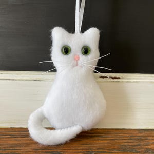 May include: White felt cat ornament with green eyes and a pink nose. The cat is sitting with its tail curled around its body.