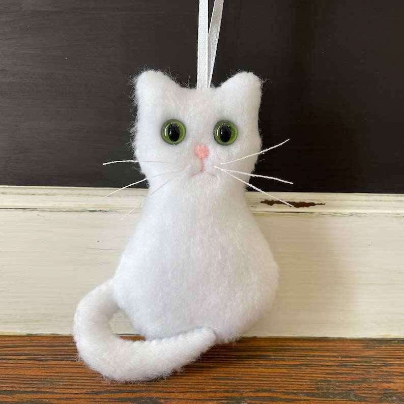 May include: White felt cat ornament with green eyes and a pink nose. The cat is sitting with its tail curled around its body.