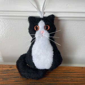 May include: A handmade black and white cat ornament with orange eyes, a pink nose, and white whiskers. The cat has a white chest and a long, curved tail. A white ribbon is attached for hanging. This cat ornament is a charming decoration.
