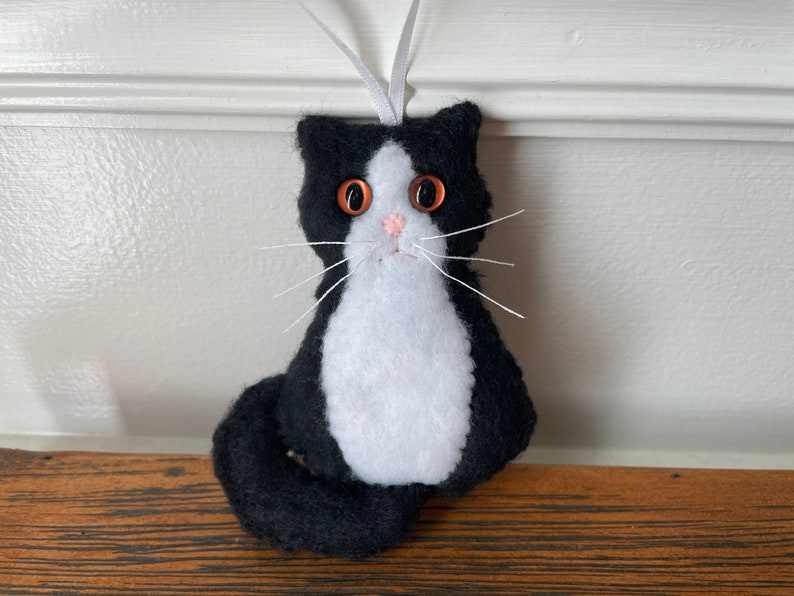May include: A handmade black and white cat ornament with orange eyes, a pink nose, and white whiskers. The cat has a white chest and a long, curved tail. A white ribbon is attached for hanging. This cat ornament is a charming decoration.