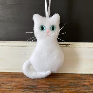 May include: White felt cat ornament with blue eyes. The cat is sitting with its tail curled around its body.