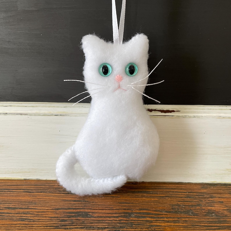 May include: White felt cat ornament with blue eyes. The cat is sitting with its tail curled around its body.