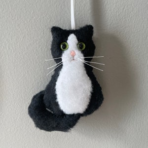 May include: A handmade felt cat ornament in black and white, with green eyes and white whiskers. The cat has a white chest and a black tail, and is hanging from a white ribbon. A cute cat decoration.