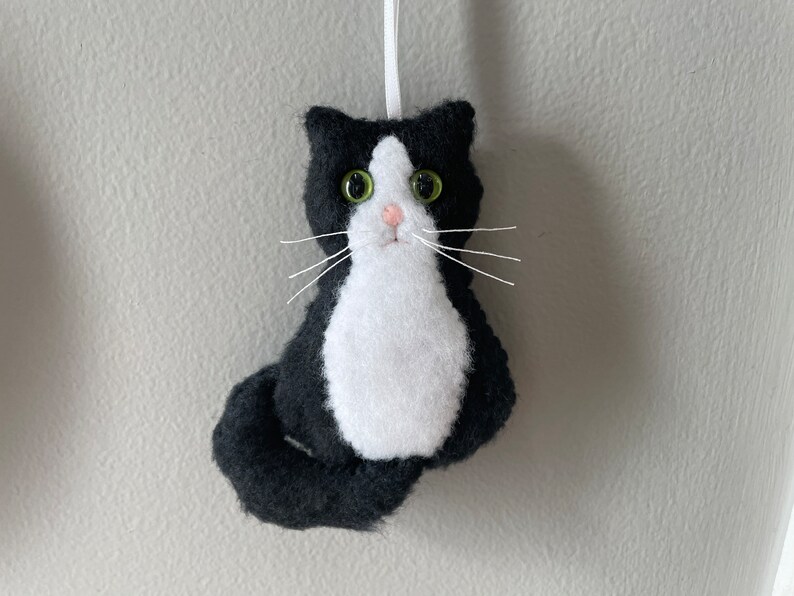 May include: A handmade felt cat ornament in black and white, with green eyes and white whiskers. The cat has a white chest and a black tail, and is hanging from a white ribbon. A cute cat decoration.