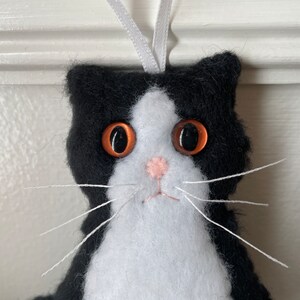 May include: A handmade felt cat ornament with a black and white pattern. The cat has large, orange eyes, a pink nose, and white whiskers. A white ribbon is attached for hanging. This cat ornament is a charming decoration.