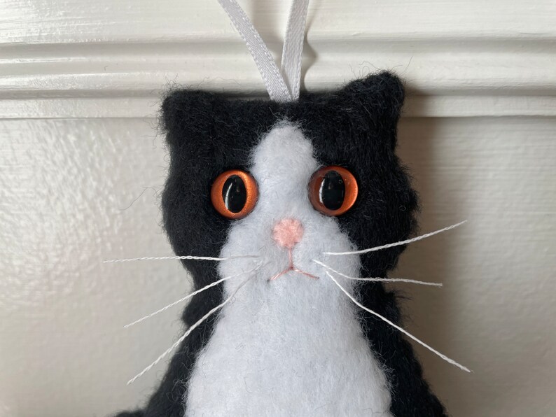 May include: A handmade felt cat ornament with a black and white pattern. The cat has large, orange eyes, a pink nose, and white whiskers. A white ribbon is attached for hanging. This cat ornament is a charming decoration.