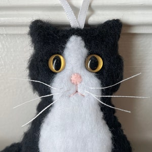 May include: A handmade black and white cat ornament with a white ribbon for hanging. The cat has large golden eyes, a pink nose, and white whiskers. The ornament is made of felt.