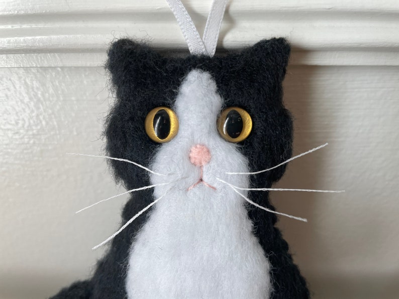 May include: A handmade black and white cat ornament with a white ribbon for hanging. The cat has large golden eyes, a pink nose, and white whiskers. The ornament is made of felt.