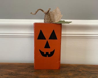 May include: A wooden, orange jack-o'-lantern decoration with a carved face and a burlap and twine stem. The jack-o'-lantern has a smiling face with triangular eyes and a jagged mouth. The stem is made of twisted twine and burlap.
