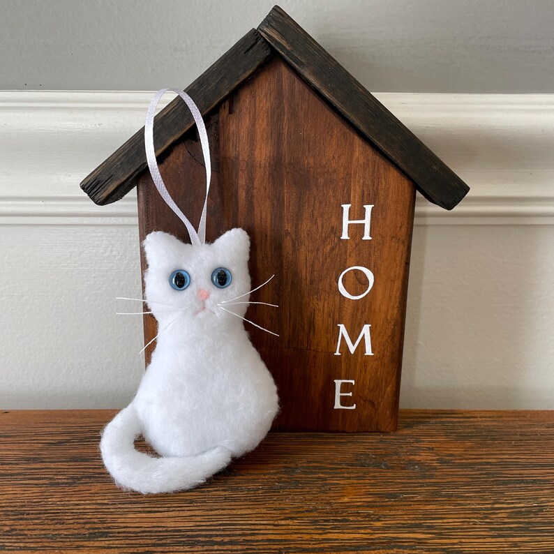 May include: White plush cat ornament with blue eyes and a pink nose. The ornament is hanging from a white ribbon and is in front of a wooden sign that says "HOME".