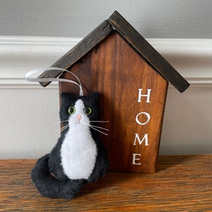 May include: A handmade felt cat ornament in black and white, with green eyes and white whiskers, hangs in front of a wooden house-shaped sign. The sign is stained brown with the white word "HOME" printed on it. A white ribbon is attached to the ornament.