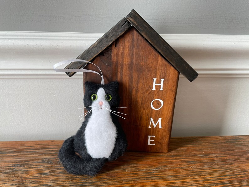 May include: A handmade felt cat ornament in black and white, with green eyes and white whiskers, hangs in front of a wooden house-shaped sign. The sign is stained brown with the white word "HOME" printed on it. A white ribbon is attached to the ornament.