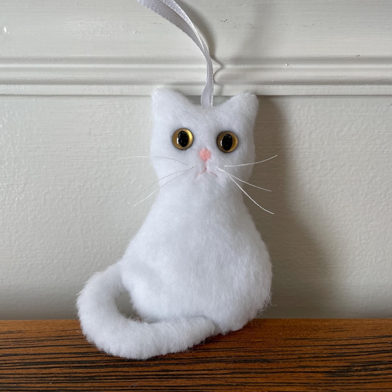 May include: White plush cat ornament with yellow eyes and a pink nose. The cat is sitting with its tail curled around its body.