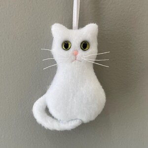 May include: White felt cat ornament with green eyes and a pink nose. The cat is sitting with its tail curled around its body.