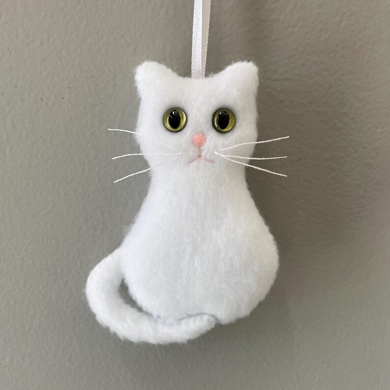 May include: White felt cat ornament with green eyes and a pink nose. The cat is sitting with its tail curled around its body.