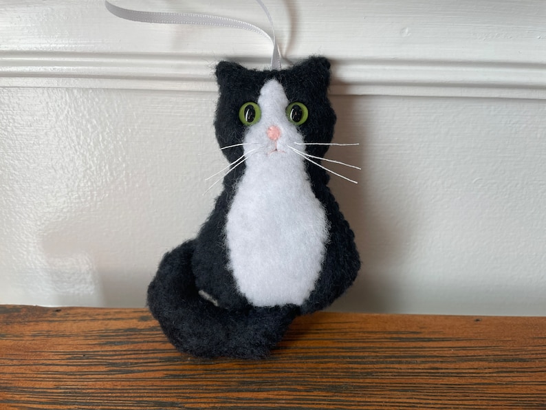 May include: A handmade felt cat ornament in black and white, with green eyes and white whiskers. The cat has a white chest and face, with a black body and tail. A gray ribbon is attached for hanging.