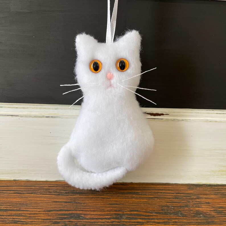 May include: White plush cat ornament with orange eyes and a pink nose. The cat is sitting with its tail curled around its body.