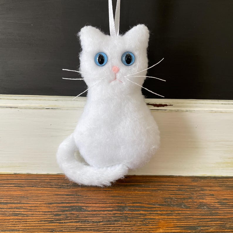 May include: White plush cat ornament with blue eyes. The cat is sitting with its tail curled around its body.