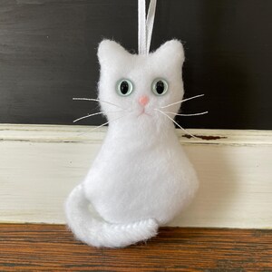 May include: White plush cat ornament with blue eyes and a pink nose. The cat has a long tail and is hanging from a white string.