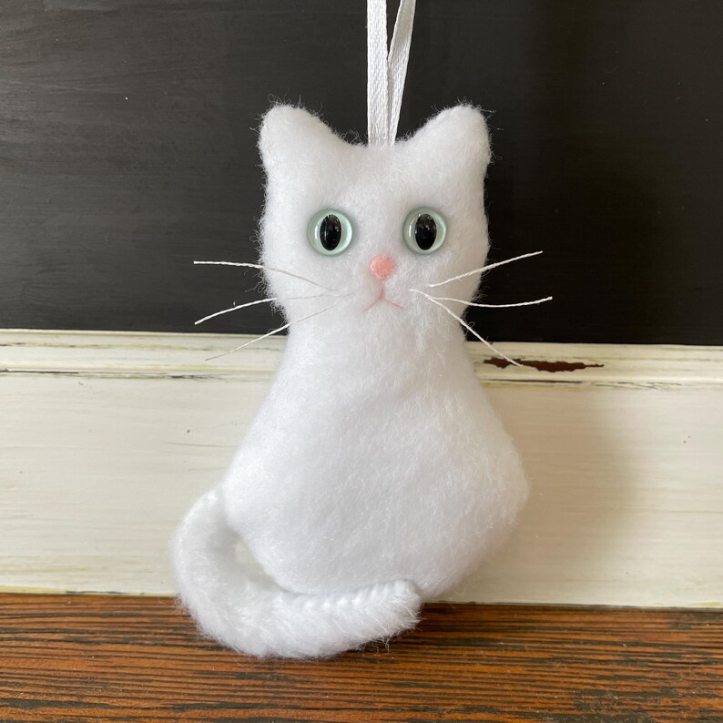 May include: White plush cat ornament with blue eyes and a pink nose. The cat has a long tail and is hanging from a white string.