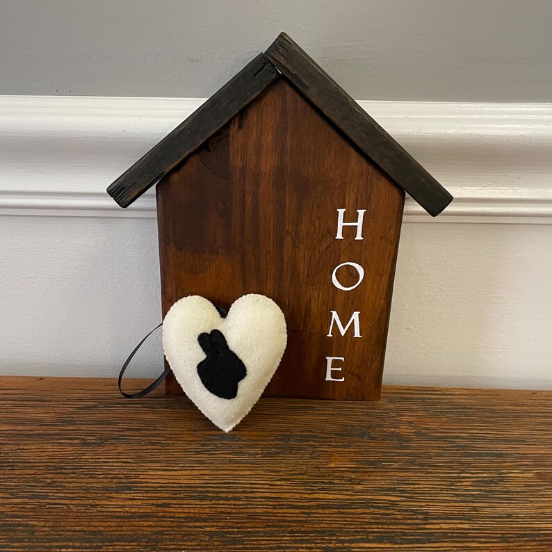 May include: A wooden house-shaped sign with the word "HOME" painted in white on the front. A white felt heart with a black bunny silhouette is hanging from the sign.