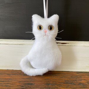 May include: White plush cat ornament with yellow eyes and a pink nose. The cat is sitting with its tail curled around its body.