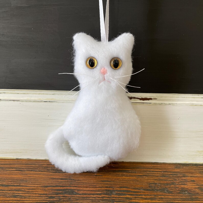May include: White plush cat ornament with yellow eyes and a pink nose. The cat is sitting with its tail curled around its body.
