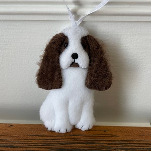 May include: A handmade felt dog ornament in white and brown. The dog has floppy brown ears, a black nose, and is sitting upright. A white ribbon is attached for hanging. The ornament is a charming decorative item.