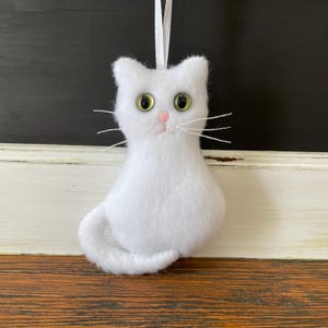 May include: White felt cat ornament with green eyes and pink nose. The cat is sitting with its tail curled around its body.
