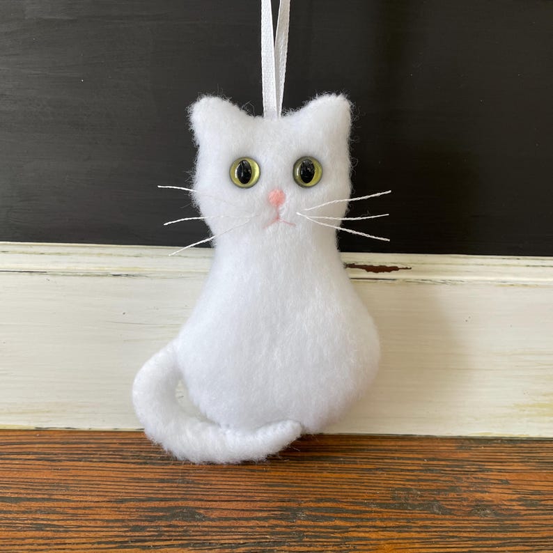 May include: White felt cat ornament with green eyes and pink nose. The cat is sitting with its tail curled around its body.