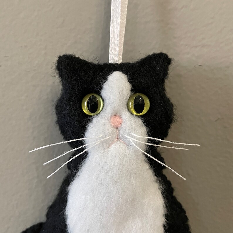 May include: A handmade felt cat ornament with a black and white tuxedo pattern. The cat has large, yellow-green eyes, a pink nose, and white whiskers. A white ribbon is attached for hanging.