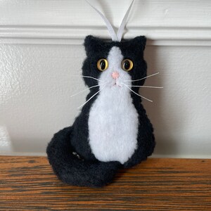 May include: A handmade black and white felt cat ornament with a white ribbon for hanging. The cat has gold button eyes, a pink nose, and white whiskers. The cat's body is black with a white chest and belly. A cute cat decoration.