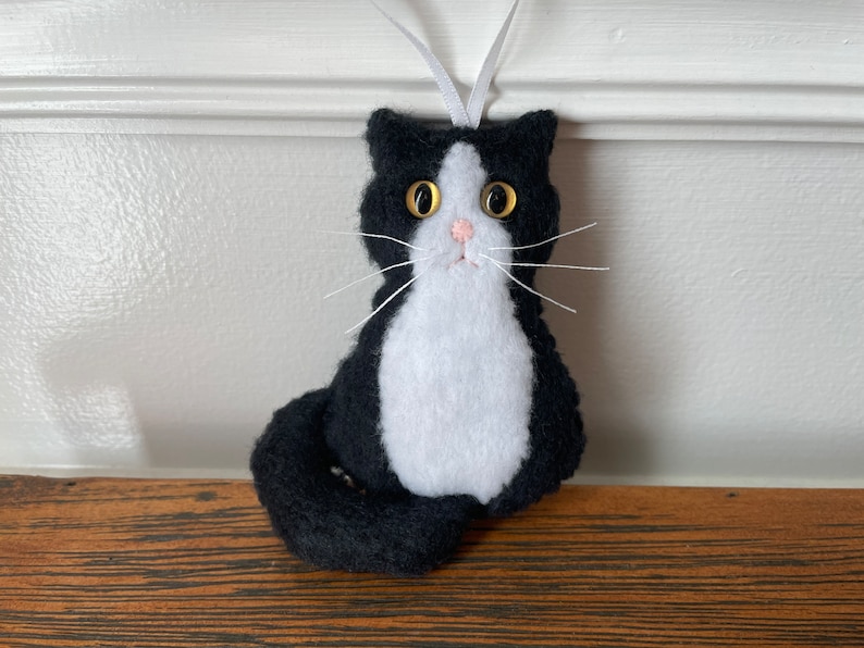 May include: A handmade black and white felt cat ornament with a white ribbon for hanging. The cat has gold button eyes, a pink nose, and white whiskers. The cat's body is black with a white chest and belly. A cute cat decoration.