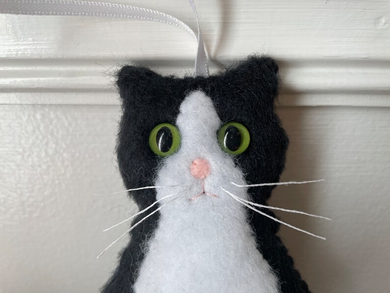May include: A handmade felt cat ornament with a black and white tuxedo pattern. The cat has large green button eyes, a pink nose, and white whiskers. A gray ribbon is attached for hanging. Cat-themed decor.