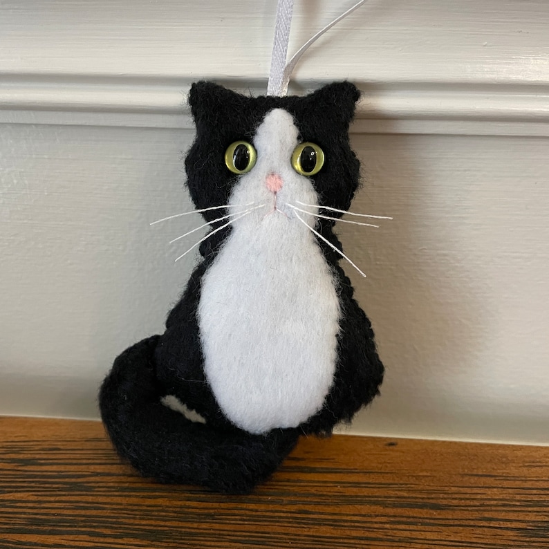 May include: A handmade black and white cat ornament with a white ribbon for hanging. The cat has large, yellow eyes, a pink nose, and white whiskers. The ornament is made of felt and is designed to look like a tuxedo cat.