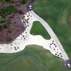 May include: Aerial view of a golf course with a winding paved road and a golf cart. The course features green grass, white sand traps, and patches of brown vegetation. The overall scene is bright and sunny.