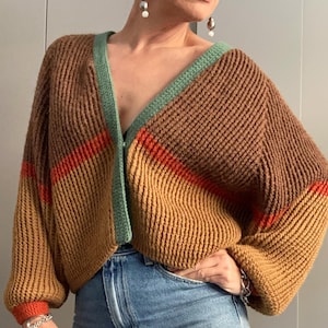 Può includere: Un cardigan lavorato a maglia marrone, arancione e verde con scollo a V. Il cardigan presenta una trama a coste, strisce arancioni sulle maniche e un bordo verde. Le maniche sono lunghe e la vestibilità è ampia. Un capo caldo e confortevole.