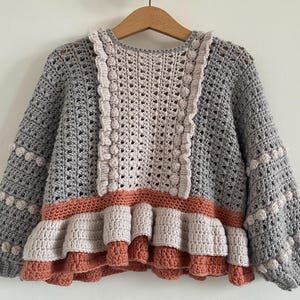 May include: A handmade crochet sweater in shades of gray, beige, and coral. The sweater features a textured stitch pattern, ruffled trim, and decorative bobbles on the sleeves. The garment is displayed on a wooden hanger against a white background.