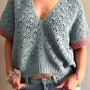 May include: A light blue crochet top with a V-neck and short sleeves. The sleeves have a pink trim. The top has an open, lacy design. The person is wearing blue jeans and a silver bracelet.