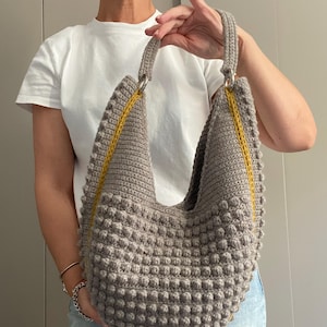May include: A gray, handmade crochet bag with a textured, bobble stitch design. The bag features a yellow accent stripe and a handle attached with silver-colored rings. The bag is held up by a person.
