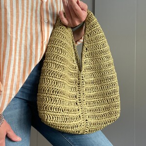 May include: A green crocheted tote bag with a wide, open top and a single strap. The bag is made of a woven material and has a slightly textured surface.
