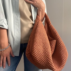 Può includere: Una borsa tote all'uncinetto strutturata color ruggine. La borsa ha una forma arrotondata e una singola tasca. La borsa è tenuta da una persona che indossa una camicia azzurra abbottonata, un top beige e jeans.