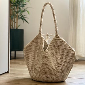 May include: A beige crochet tote bag with long handles. The bag has a wide, open top and a rounded bottom. The handles are attached to the top of the bag and are long enough to be worn over the shoulder. The bag is made of a textured yarn.