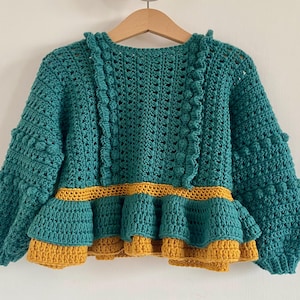 Puede incluir: Suéter de ganchillo turquesa y amarillo mostaza con dobladillo con volantes. El suéter hecho a mano presenta un patrón de puntadas texturizadas, mangas largas y cuello redondo. El suéter se muestra en una percha de madera sobre un fondo blanco.