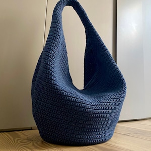 May include: A large, dark blue crocheted tote bag with a single strap. The bag is made of a thick, textured yarn and has a slouchy, casual shape.