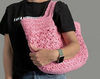 May include: A pink crocheted tote bag with a flower pattern. The bag has two straps and is being held by a person wearing a black t-shirt and blue jeans.